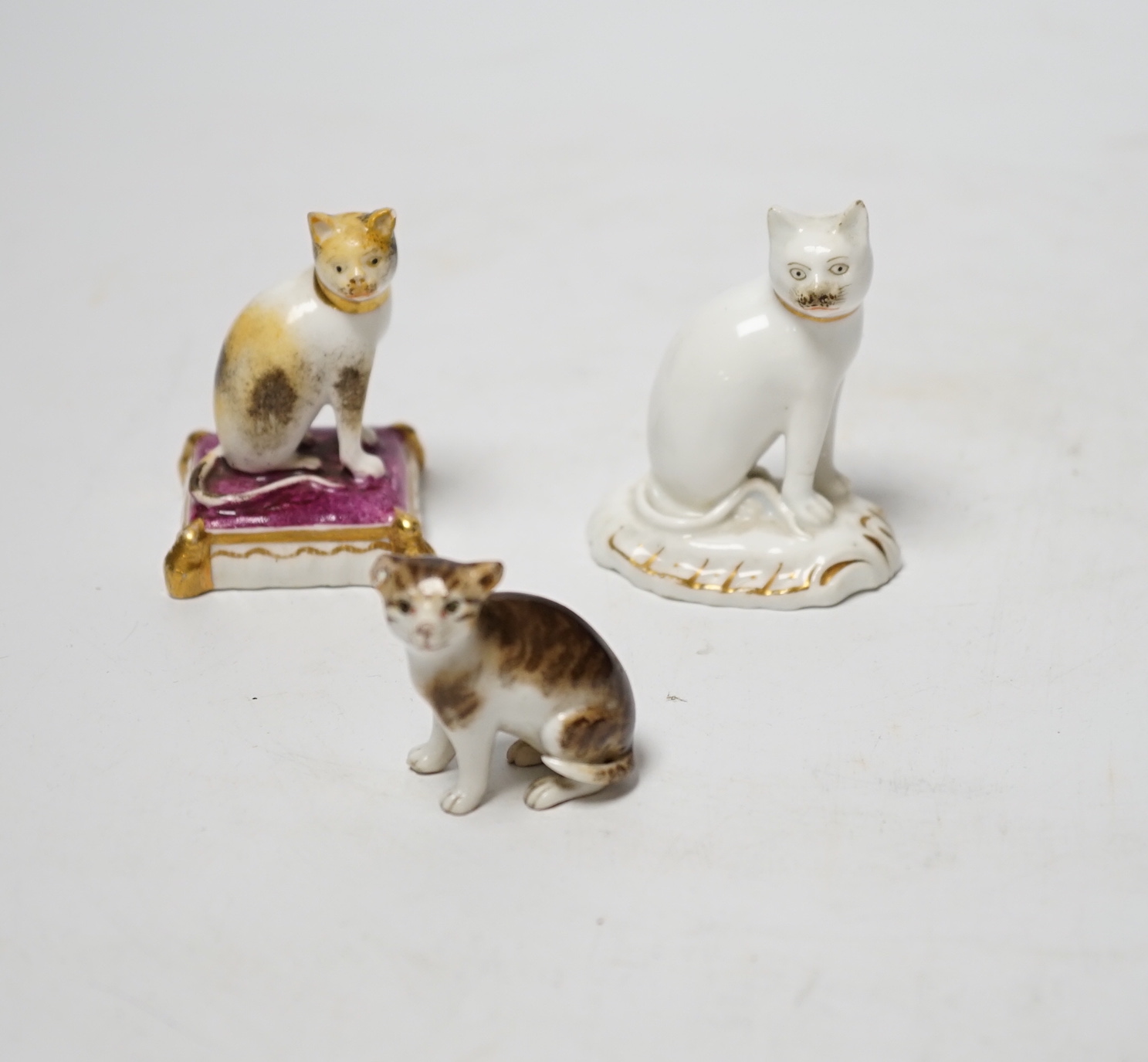 Two Rockingham porcelain models of seated cats, c.1830, the cat seated on a maroon cushion with impressed Rockingham Mark and incised model number 17, 5.3cm high, the white and gilt example with incised model number 11,